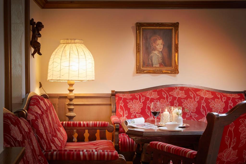 Siltā un mājīgā istabā ir sarkanas krāsas dīvāns un krēsli, dekorēti ar ziedu rakstu. Uz galda atrodas vīna glāze un ēdieni, bet sienā piekārts glezna ar bērna portretu. Lampa ar auduma abažūru piešķir gaisotnei intimitāti.
