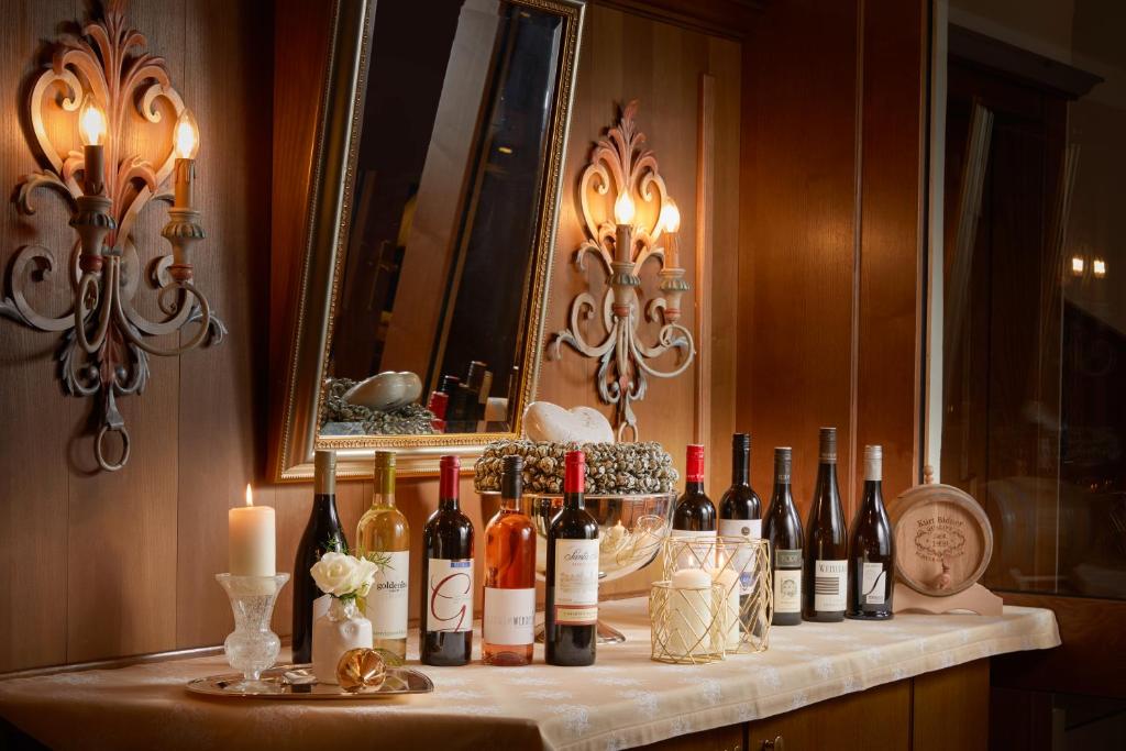 Eleganta galda kompozīcija ar dažādām vīna pudelēm, sveci un ziediem, radot viesmīlīgu atmosfēru. Interjers ar koka apdari un spoguli, kas piešķir siltumu un mājīgumu.