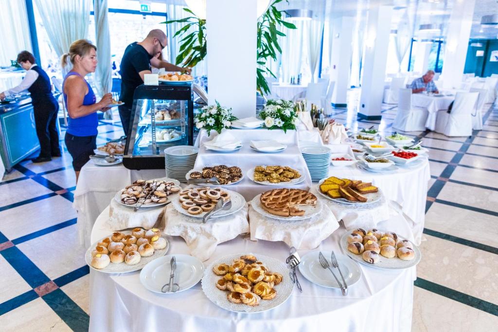 Bagātīgs bufetes galds ar dažādām konditorejas izstrādājumiem un uzkodām, kas pasniegts modernā restorānā. Galds rotāts ar ziediem un piedāvā plašu izvēli gardumu un saldumu.