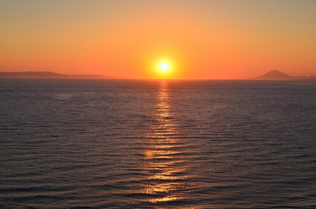 Saulriets vir jūru, radot siltas oranžas un dzeltenas nokrāsas, kas atspoguļojas ūdenī. Skats uz horizontu ar kalniem fona.