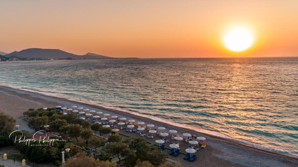 Saulriets virs klusa jūras pludmales, kur redzami sauļošanās krēsli un saulessargi. Fons veido kalni un mierīga jūra, radot relaksējošu atmosfēru.