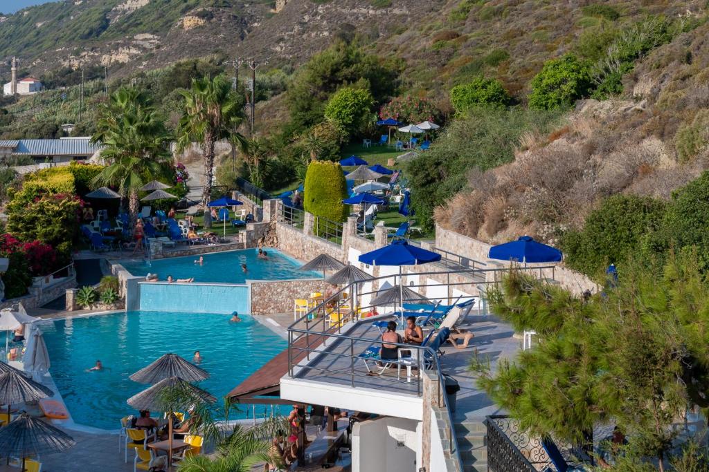 Peldbaseins ar sauļošanās krēsliem un palmiem, apkārtne ar ziediem un kalnu fona. Apmeklētāji bauda atpūtu un sauļošanos.