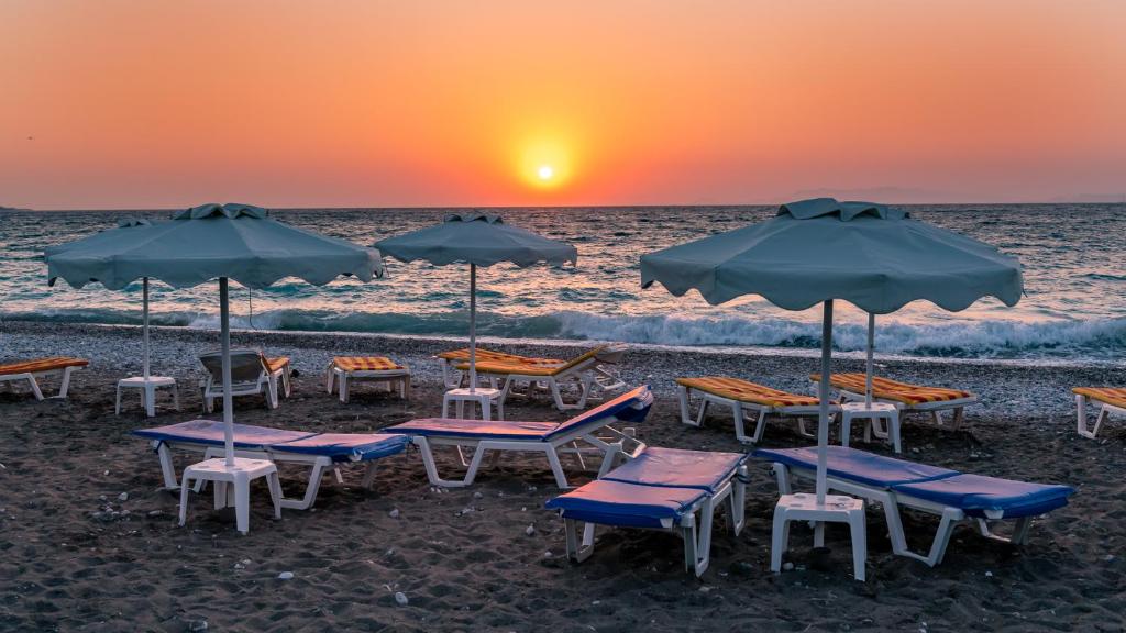 Pludmales ainava ar saulrietu, kur redzami sauļošanās krēsli un lietussargi. Jūras viļņi lēnām plūst krastā, radot mierīgu atmosfēru.