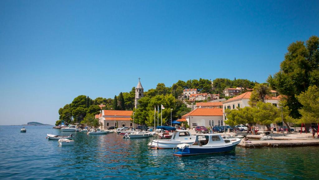 Skaists skats uz ostu ar laivām un krāšņām ēkām, kas atrodas blakus ūdenim, ko ieskauj zaļi koki un skaidra debess.