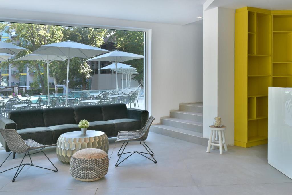 Mūsdienīgs dzīvojamais interjers ar dīvānu, galdu un krēsliem, kas vērsti pret lielu logu, caur kuru redzams peldbaseins un saulessargi. Dzeltena grāmatu plaukts un kāpnes pievieno mūsdienīgu akcentu.