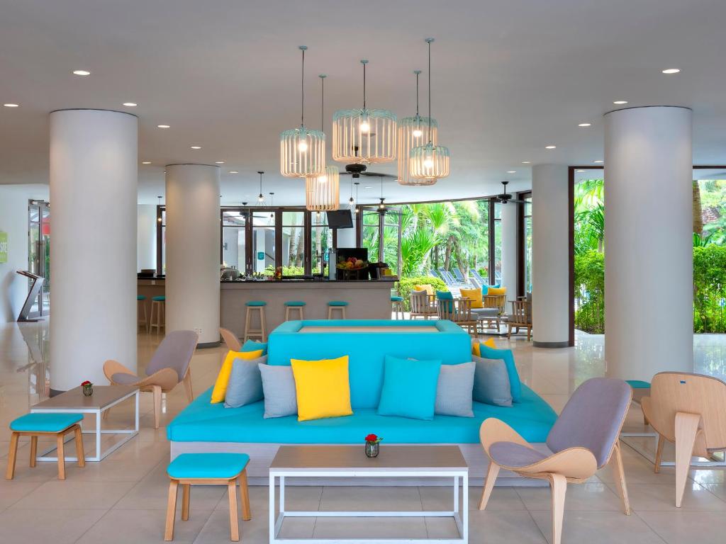 Mūsdienīgs viesnīcas interjers ar spilgtiem dīvāniem un ērtām krēsliem, kas rada relaksējošu atmosfēru. Telpā ir daudz dabiskās gaismas un zaļa augšana, kas piešķir svaigumu.