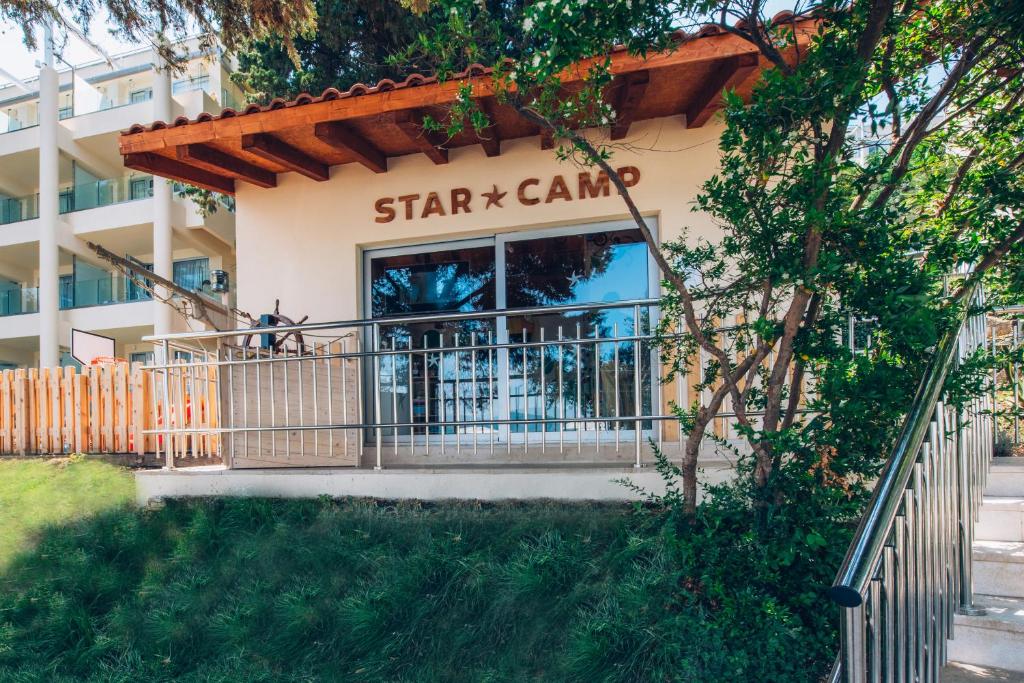 Bērnu nometnes "STAR CAMP" ēka ar koka jumtu, kas atrodas zaļā vidē, blakus modernām viesnīcām.