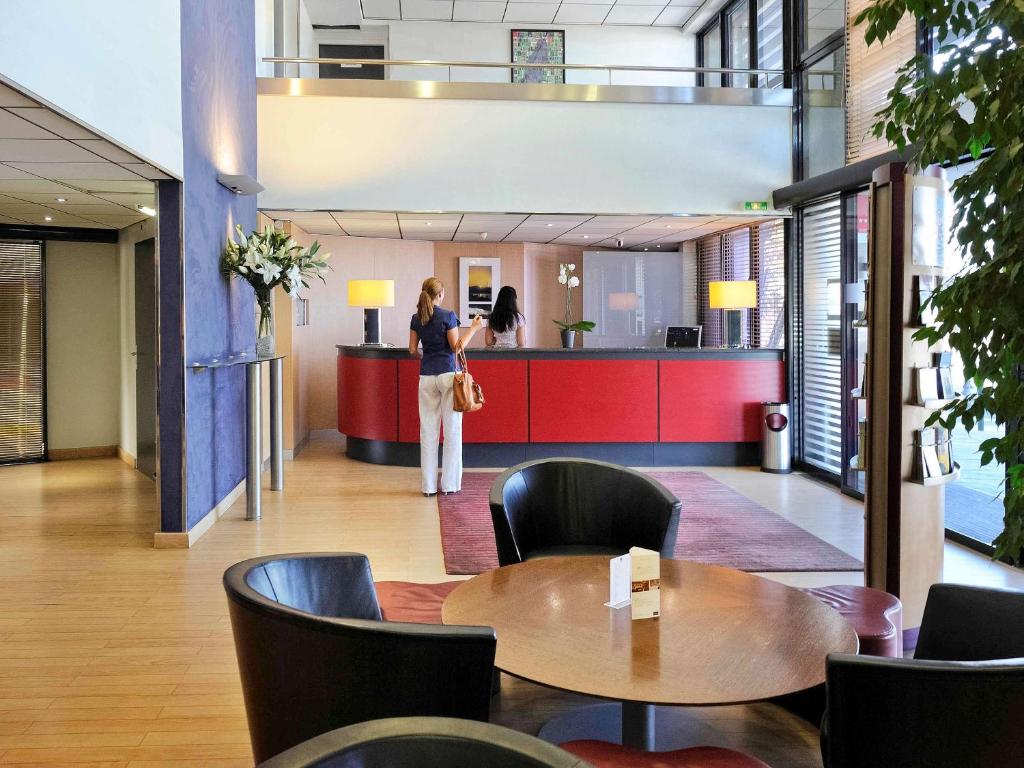 Mūsdienīgs viesnīcas reģistratūras interjers ar sarkanu pieņemšanas letni un ērtām melnām krēslām, kur viesi gaida apkalpošanu. Dabiska gaisma plūst cauri lielajiem logiem, radot siltu un aicinošu atmosfēru.