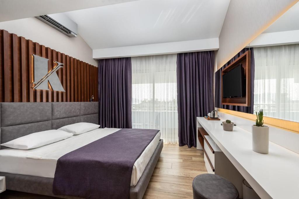 Mūsdienīga viesnīcas numura iekārtojums ar pelēku gultu, tumši violetām aizkariem un elegantu rakstāmgaldu. Telpa ir gaiša un gaumīga, nodrošinot komfortablu atpūtu.