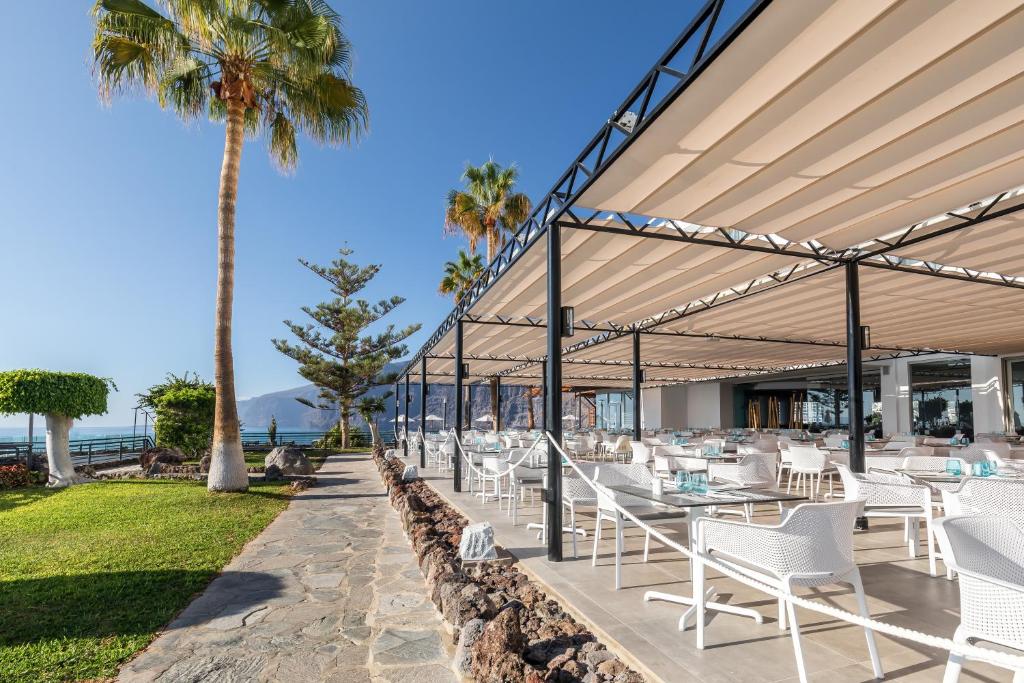 Moderns restorāns ar terasi, kas piedāvā skatu uz jūru, apkārt palmām un zaļiem krūmiem. Atvērts un saulains ēdināšanas laukums aicina viesus baudīt ēdienus ārā.