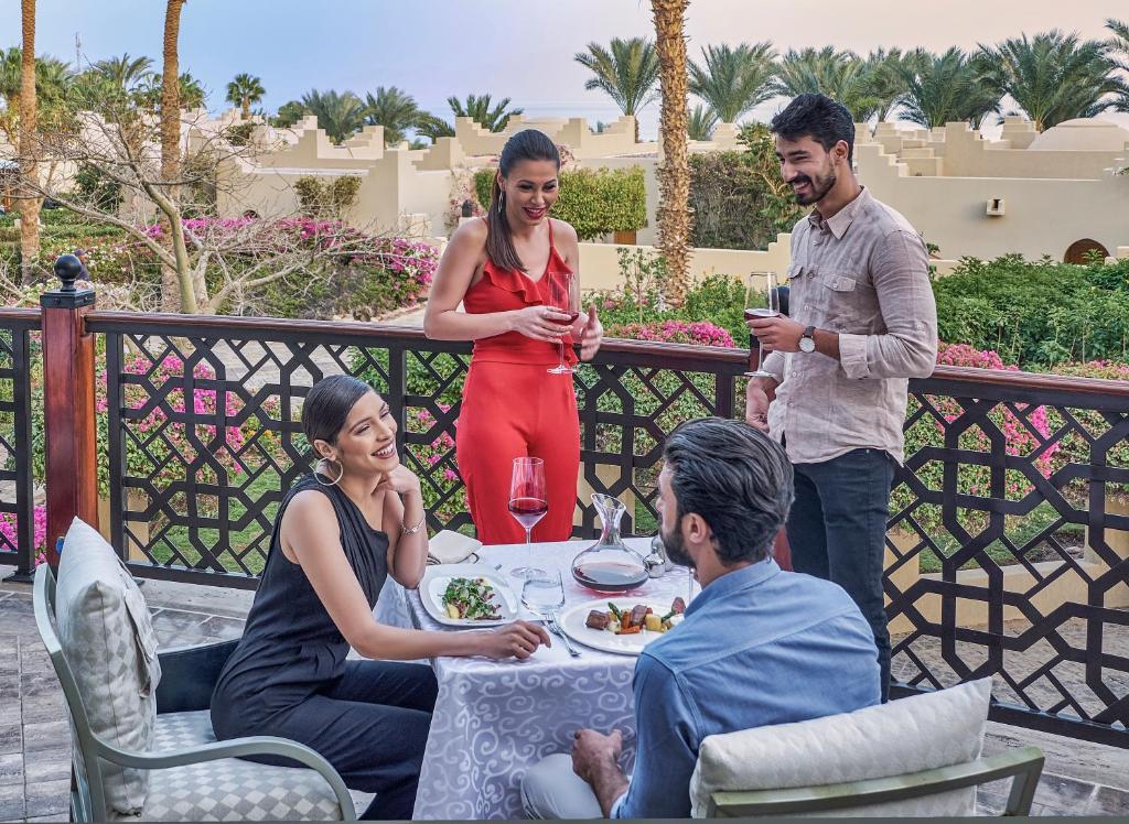 Četri cilvēki svin kopā pie galda uz terases, baudot gardus ēdienus un dzērienus, ar krāšņiem ziediem un palmām fona.