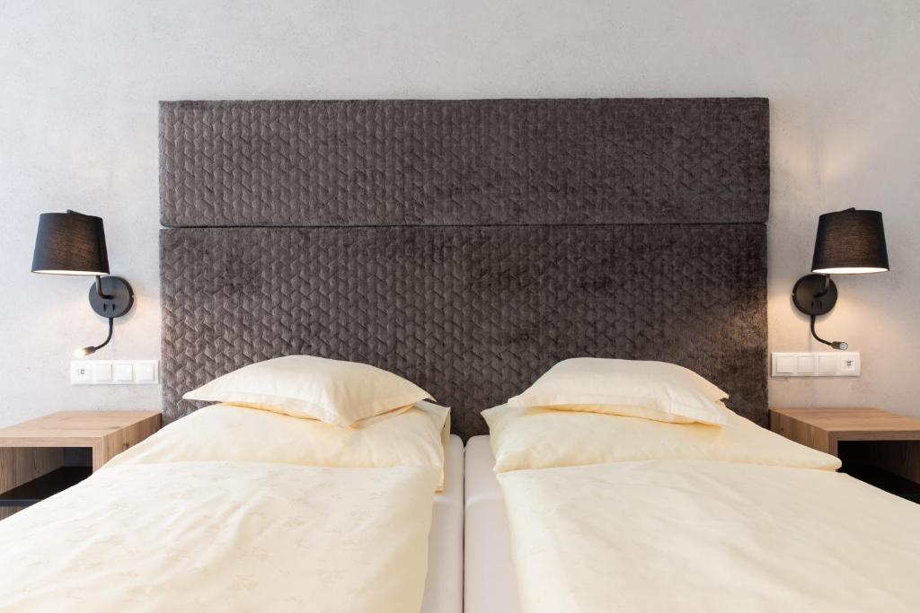 Mūsdienīgs guļamistabas interjers ar diviem gultām, pelēku galvgalvi un melnu lampu katrā pusē. Gultas ir klātas ar gaiši dzeltenām gultas veļām.