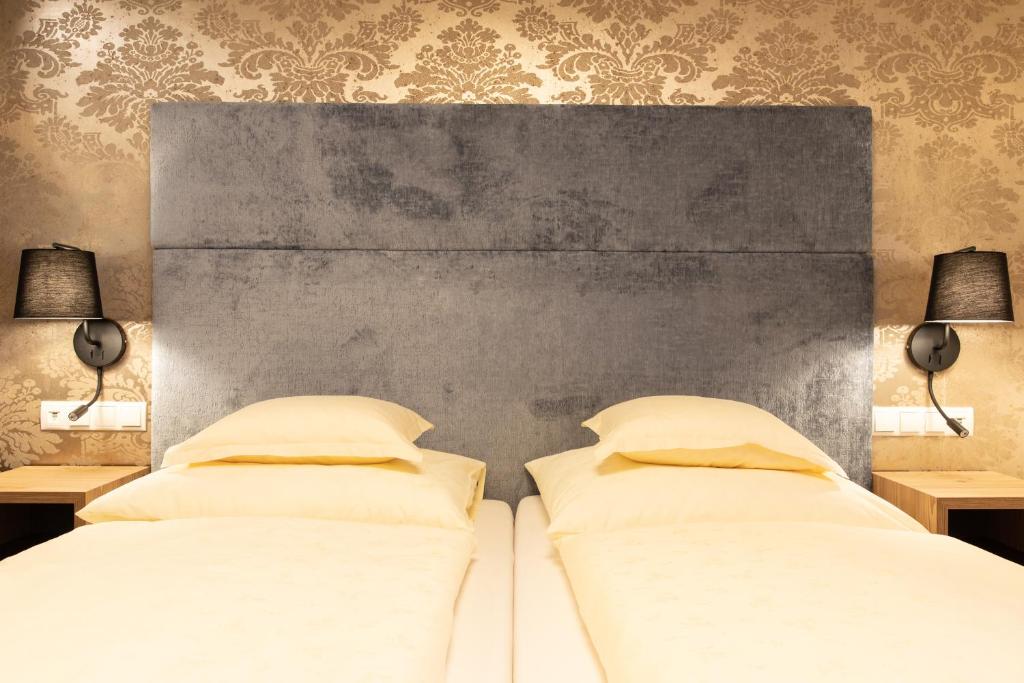 Eleganta guļamistaba ar mīkstu pelēku gultas galvu un dzeltenām gultām, fona tapetes ar dekoratīvu rakstu. Apgaismojums no sienas lampām rada siltu atmosfēru.