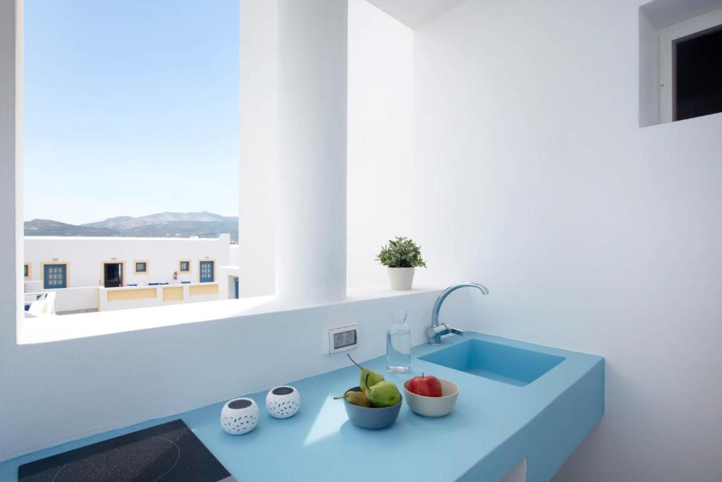 Mūsdienīgs un gaišs virtuves stūris ar zilu virsmu, uz kuras atrodas augļu bļoda un ūdens karafe. Skats no loga uz kalniem un baltas ēkas.