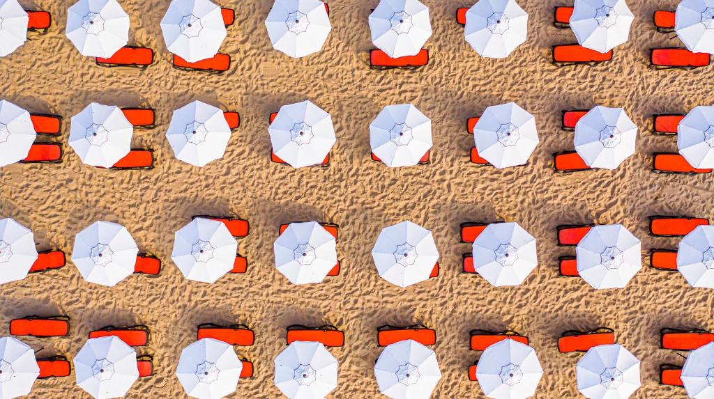 Pludmales sauļošanās krēsli ar baltām saulessargiem, sakārtoti organizētās rindās smiltīs. Ideāla vieta atpūtai un relaksācijai.