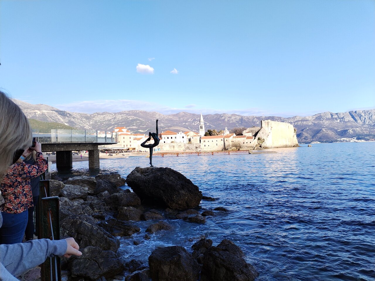Skatu punkts uz jūru ar skulptūru, kas attēlo dejotāju uz akmens. Fona ainavā redzams vēsturīgs pilsētas centrs un kalni, kas veido gleznainu apkārtni. panoramic view with beach and pools