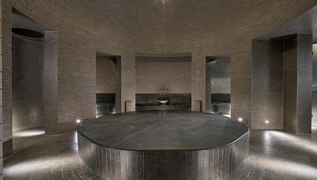 Mūsdienīga spa telpa ar apaļu akmens platformu centrā un mūra sienām, kas izveido elegantu un relaksējošu atmosfēru. Apgaismojums un ūdens elementi piešķir telpai mierīgu noskaņu.