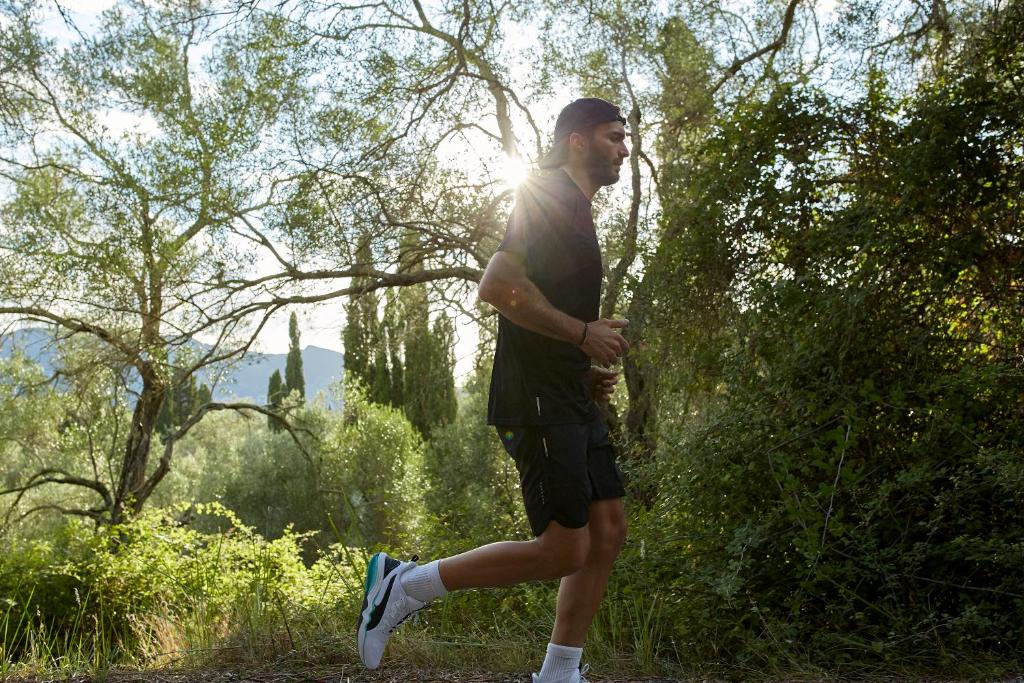 Vīrietis skrien pa meža taku, apkārt augoši koki un saules gaisma caur lapām. Viņš ir sportiskā apģērbā un izskatās enerģisks.