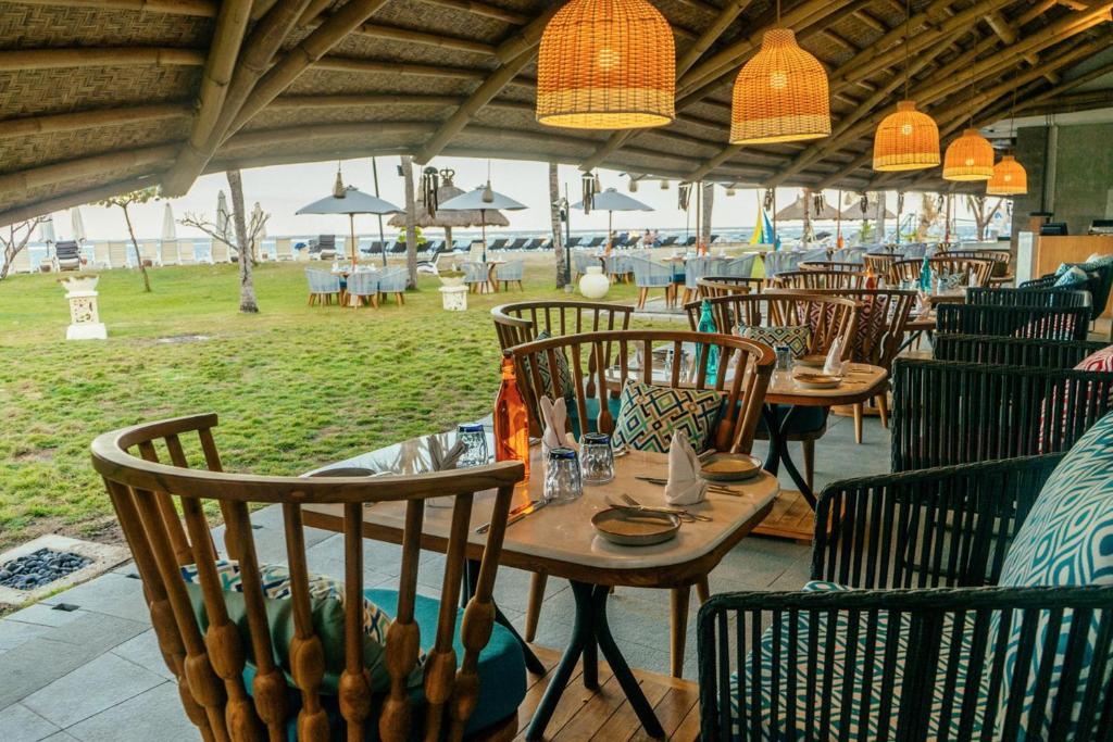 Elegantā restorāna interjers ar koka mēbelēm un rotājošiem lampām, kas piedāvā skatu uz zaļo zāli un pludmali. Rāmie krēsli un galdi ir sagatavoti vakariņām brīvā gaisotnē.