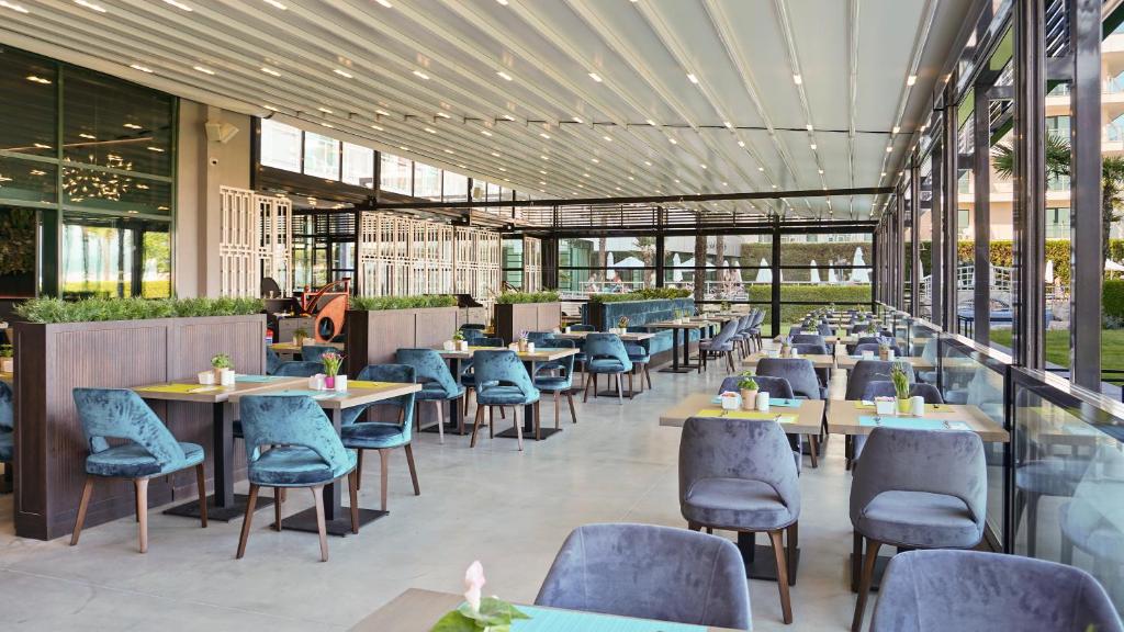 Mūsdienīgs restorāns ar plašu stiklotu terasi, kurā ir eleganti galdi un krēsli. Telpa ir gaiša un atvērtā, piedāvājot komfortablu atmosfēru ēdiena baudīšanai.