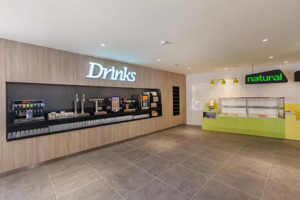 Mūsdienīga dzērienu stacija ar apgaismotu uzrakstu "Drinks" un zaļo "natural" zīmi. Interjers apvieno koka un flīžu elementus, piedāvājot dažādas dzērienu izvēles iespējas.