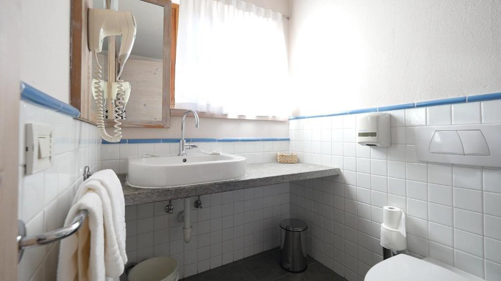 Neliels, gaišs vannas istabas interjers ar pelēku izlietni, baltiem flīzēm un zilām akcentu līnijām. Redzams arī sienas telefons un dažādi sanitārie piederumi.