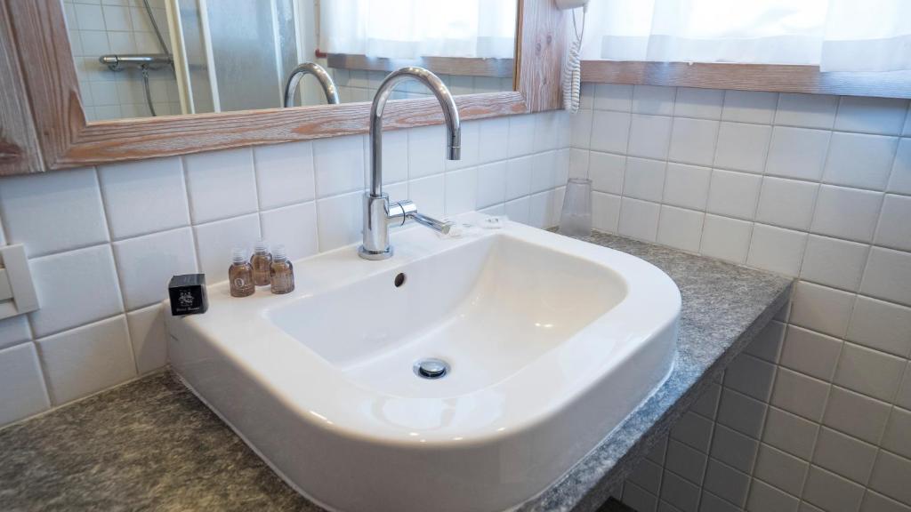 Mūsdienīgs vannas istabas izlietne ar sudraba krāsu krānu un nelielām higiēnas pudelītēm uz akmens virsmas. Fons veidots ar flīzēm un koka rāmjiem, radot siltu atmosfēru.