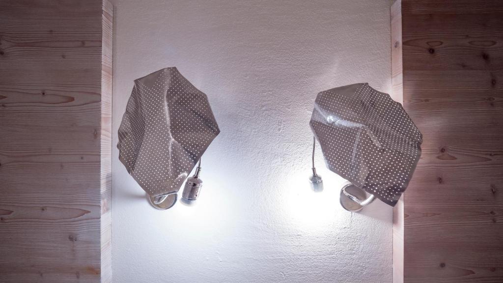 Divas modernas lampas ar punktiem, piestiprinātas pie sienas, izstaro siltu gaismu. Sienas apdare ir koka, radot mājīgu atmosfēru.