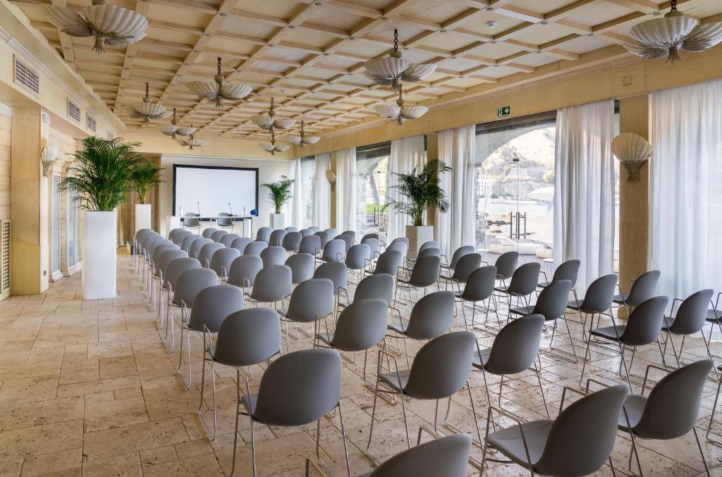 Mūsdienīga sanāksmju telpa ar rindu pelēku krēslu, lielu ekrānu un dabīgām gaismām, kas ieplūst caur logiem. Telpā ir dekoratīvi augi un elegants interjers.