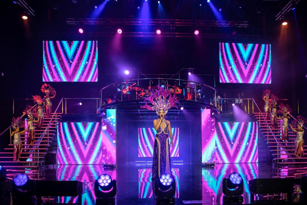 Skatuves priekšnesums ar dejotājiem un dziedātāju, kas tērpti greznos tērpos. Aiz viņiem redzami krāsaini ekrāni un spilgtas gaismas, radot dinamisku atmosfēru.