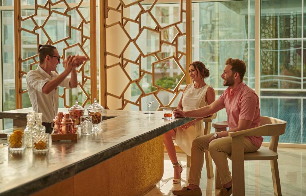 Elegants bāra skats ar bārmeni, kurš gatavo kokteiļus, un diviem apmeklētājiem, kas izbauda dzērienus. Gaismas un modernā arhitektūra rada patīkamu atmosfēru.