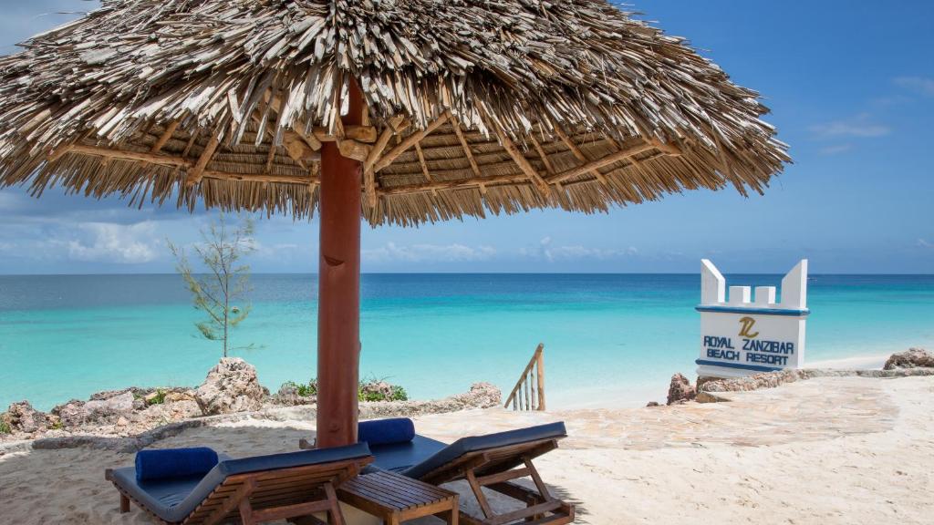 Pludmales atpūtas zona ar salmu jumtu un sauļošanās krēsliem, redzama Zanzibā, blakus Royal Zanzibar Beach Resort zīmei. Skats uz skaistu, zilganu jūru un debesīm.