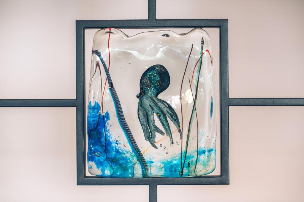 Stikla mākslas darbs ar oktopoda attēlu, kas izveidots ar krāsainiem elementiem un ūdens efektiem. Šis radošs darbs ir ievietots melnā rāmī, radot modernu un pievilcīgu izskatu.