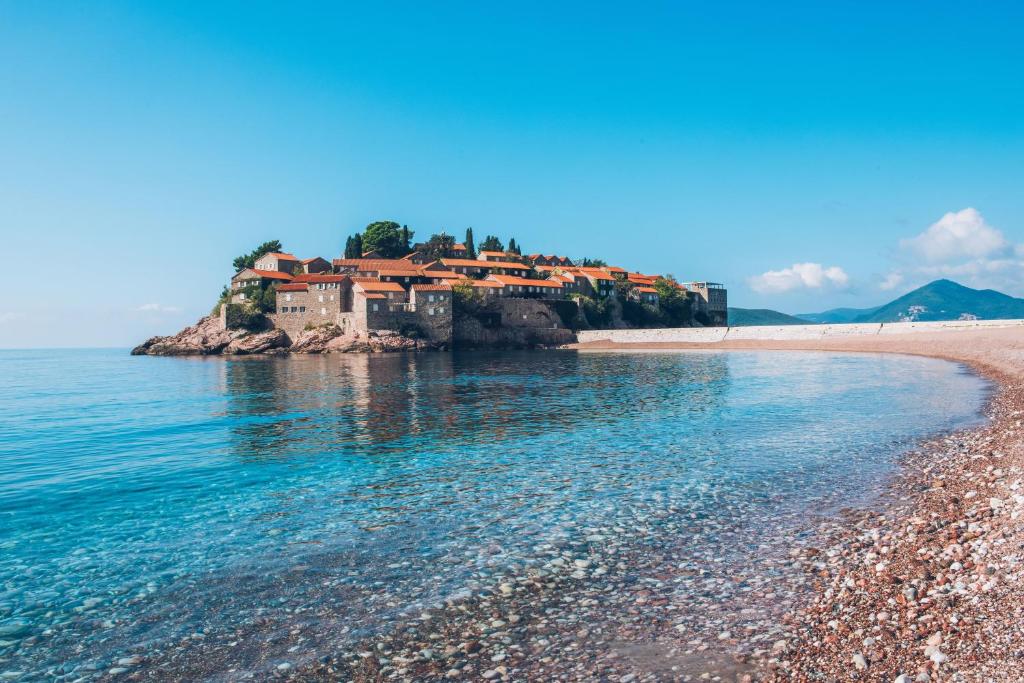 Skaista pludmale ar dzidru ūdeni un vēsturisku ēku kompleksu, kas atrodas uz salas. No attāluma redzams, ka ēkas ir ar sarkaniem jumtiem un zaļiem kokiem.