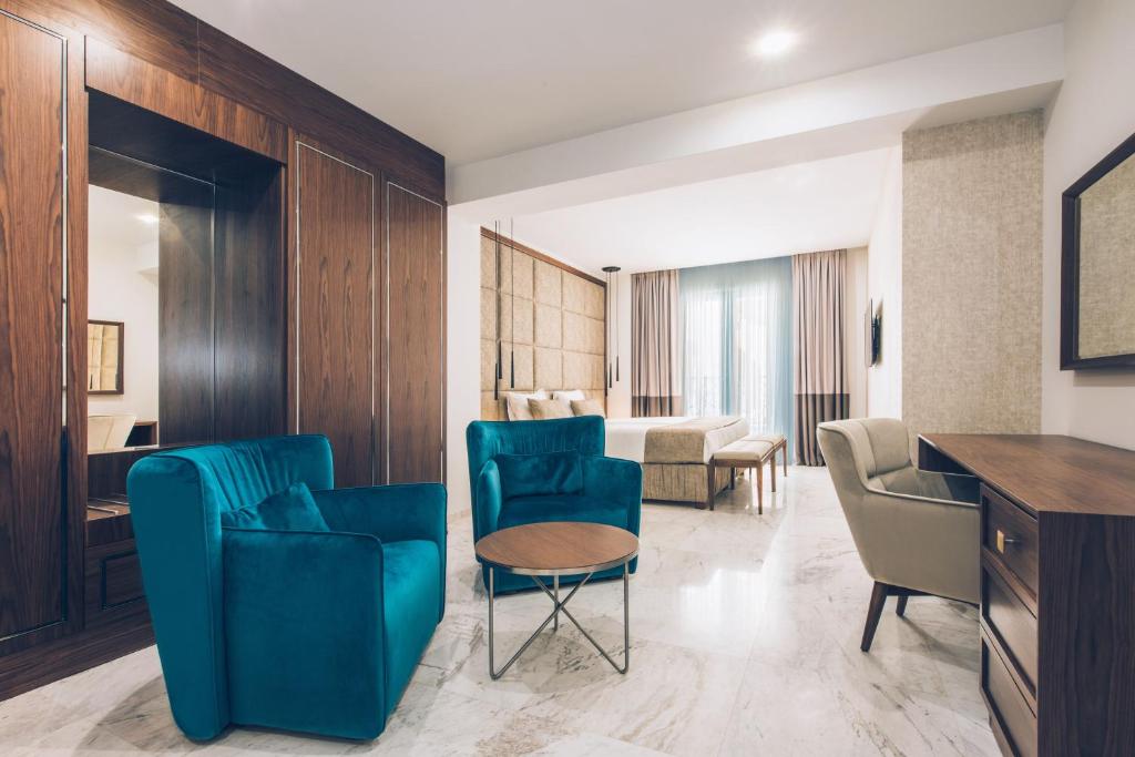 Mūsdienīga viesnīcas numura interjers ar stilīgām zilo krēslu grupām un koka akcentiem. Telpa ir gaiša un plaša, ar elegantu galdu un ērtiem gultas rāmjiem.