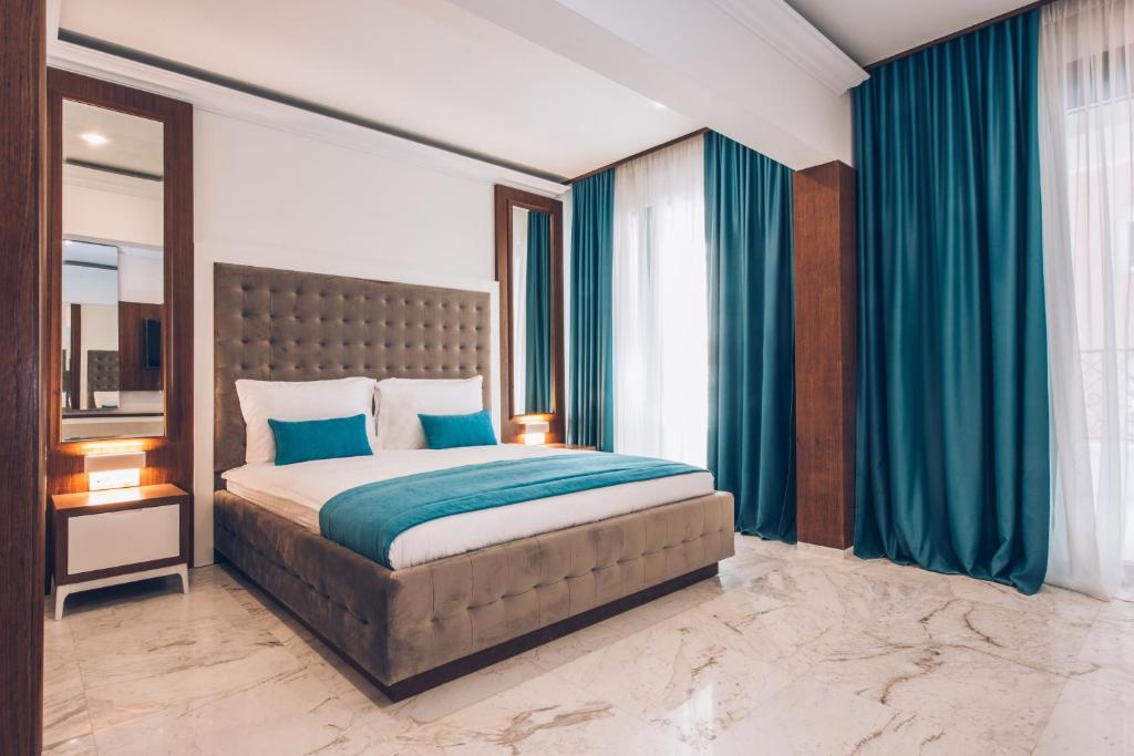 Mūsdienīgs guļamistabas interjers ar pelēku gultu un zilu gultas pārklāju, ko ieskauj eleganti zili aizkari un marmora grīda. Dabiska gaisma plūst caur logiem, radot patīkamu atmosfēru.