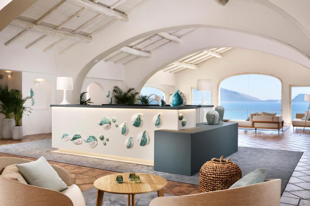 Mūsdienīgs viesnīcas reģistratūras interjers ar gaišām krāsām un jūras tematiku, kas rada relaksējošu atmosfēru. Logi piedāvā skatu uz jūru, bet telpā ir eleganti mēbeļu elementi un zaļi akcenti.