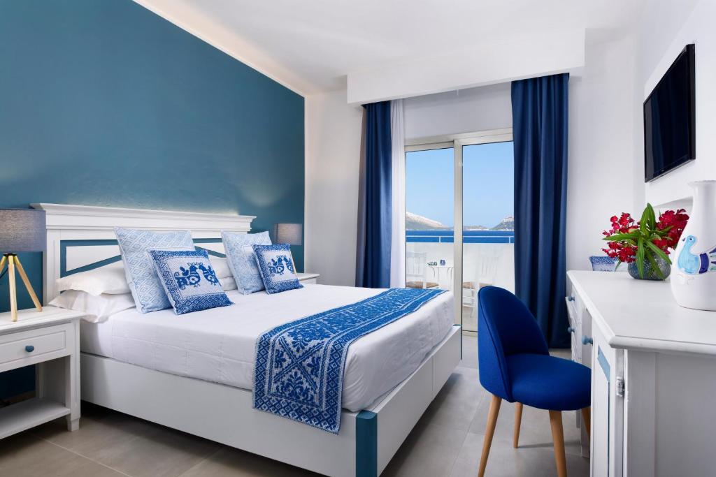 Mūsdienīga guļamistaba ar zilām sienām, baltu mēbeļu komplektu un skatu uz jūru. Interjers ir elegants, ar ziediem uz galda un televizoru pie sienas.