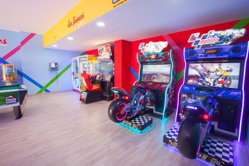 Krāsains spēļu istabas interjers ar dažādām arcade spēlēm, tostarp motociklu sacensību spēlēm un spēļu automātiem. Telpā ir gaišas krāsas un moderns dizains, radot izklaidējošu atmosfēru.