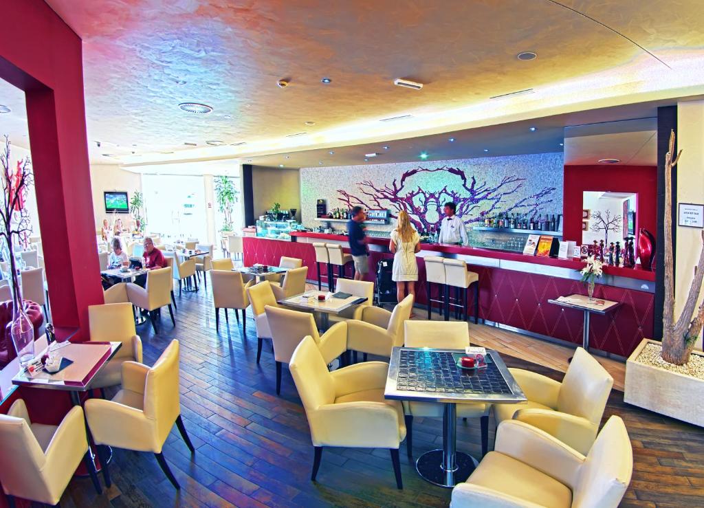 Mūsdienīgs restorāns ar elegantu interjeru, kurā ir gaišas krēsli un koka grīda, apmeklētāji bauda maltītes un dzērienus pie bāra. Restorāna siena rotāta ar māksliniecisku zaru attēlu.