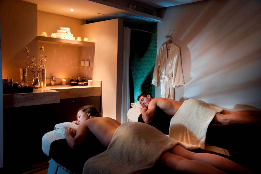 Spa procedūra pāriem relaksējošā vidē, kur divi cilvēki guļ uz masāžas galdiem, pārklāti ar dvieļiem. Telpā ir siltas gaismas un dekoratīvi elementi, kas veido mierīgu atmosfēru.