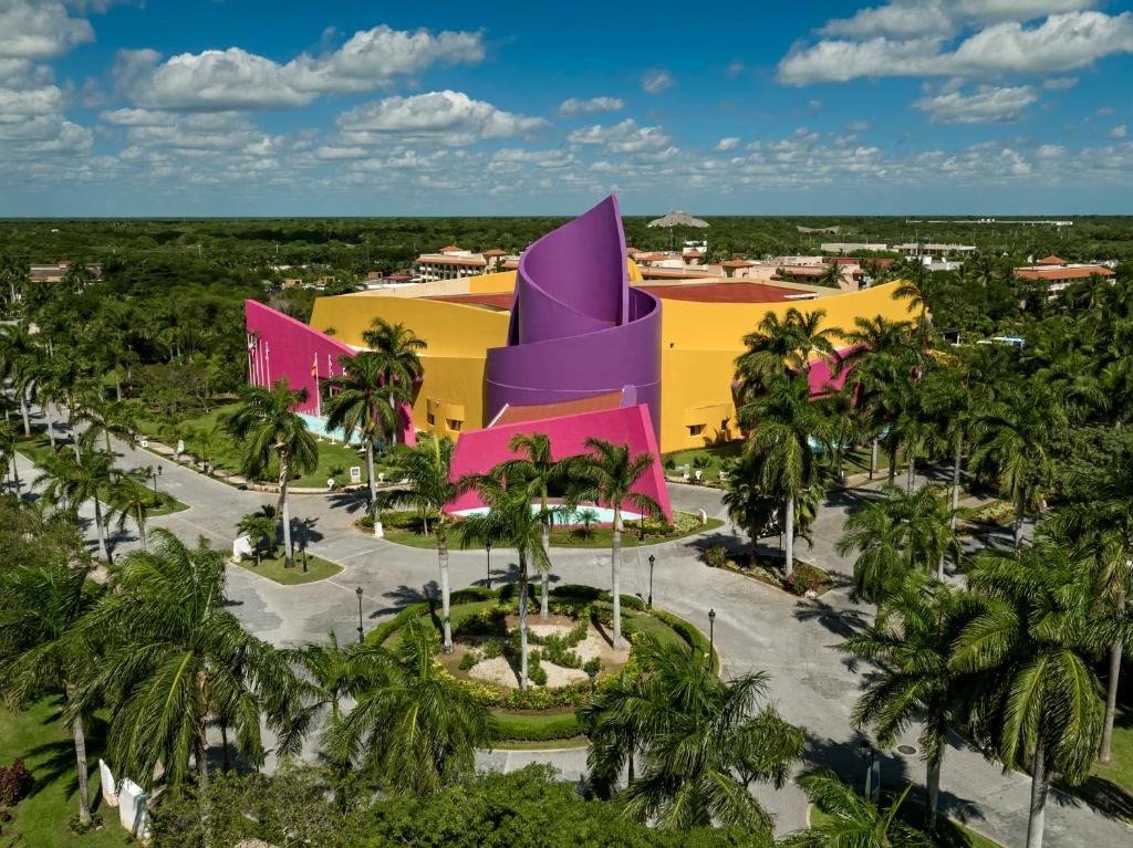 Krāsaina mūzikas un kultūras ēka ar unikālu arhitektūru, ieskauta tropisko palmu dārzā. Plašs skats uz apkārtni ar zilu debesu fonu.