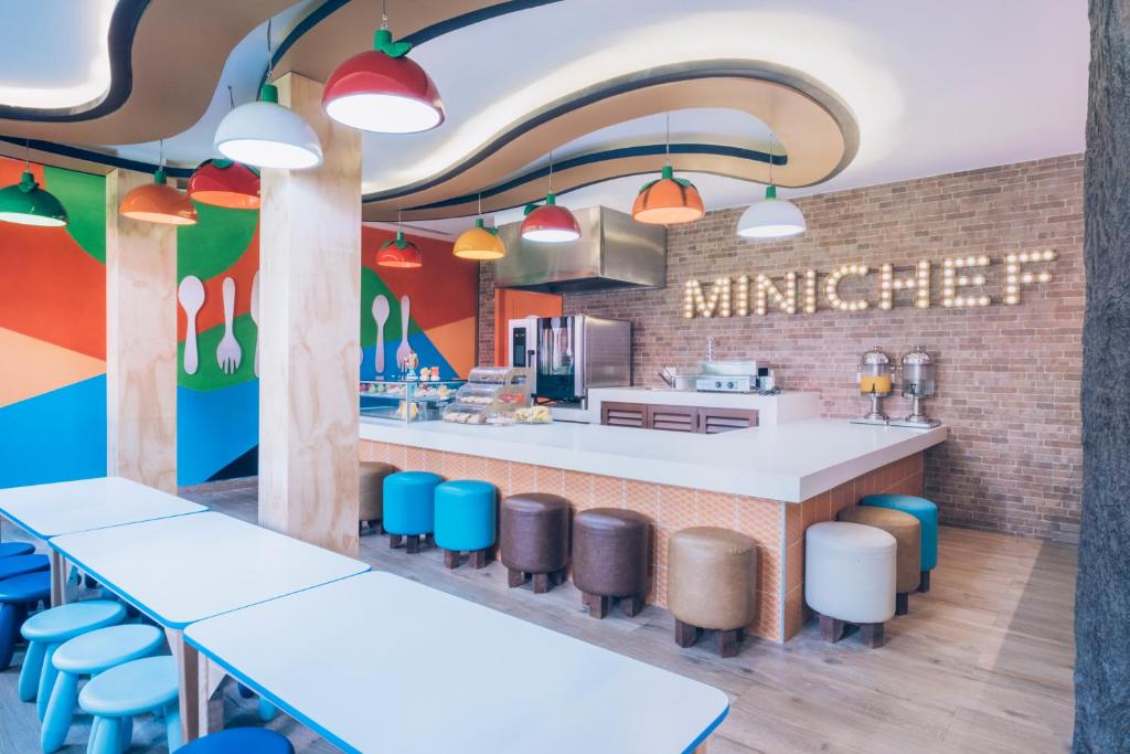Krāsaina un mūsdienīga virtuve ar zemu galdu un dažādu krāsu krēsliem, kā arī uzraksts "MINICHEF" ar gaismas diodēm uz ķieģeļu sienas. Interjers ir veidots ar radošu un jautru dizainu, kas piesaista uzmanību.