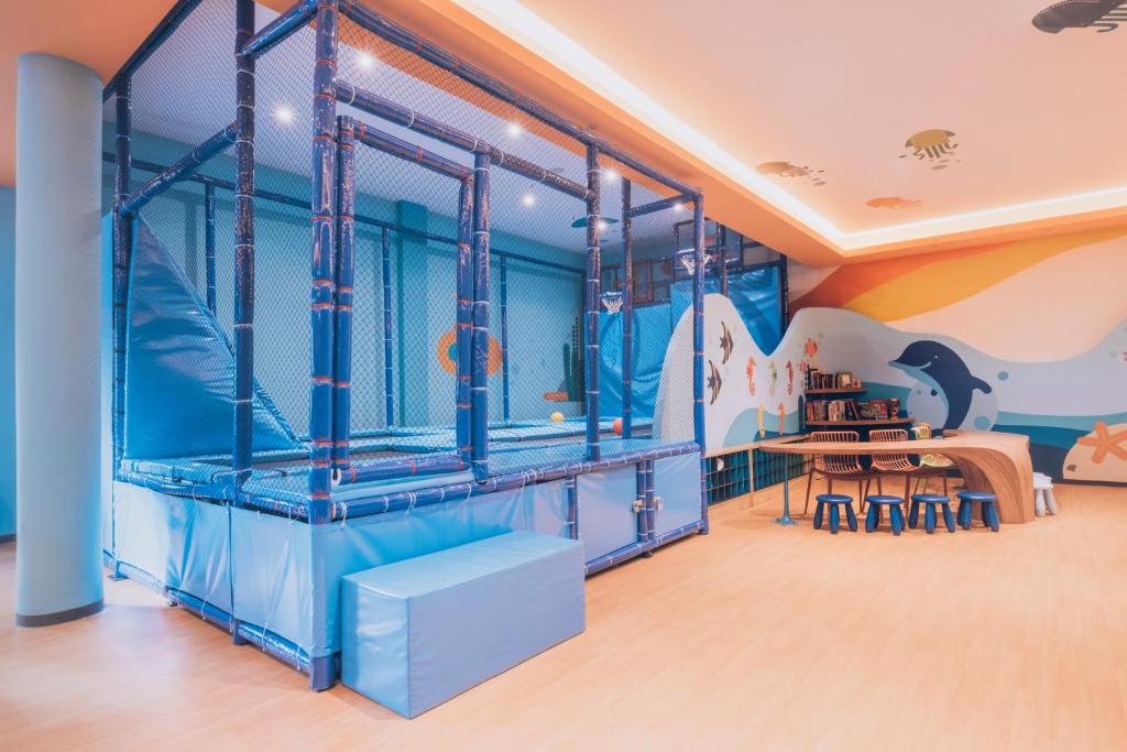 Trampolīnu zona bērnu spēļu telpā ar krāsainām sienām un rotaļu galdu. Ideāla vieta bērnu aktivitātēm un izklaidei.