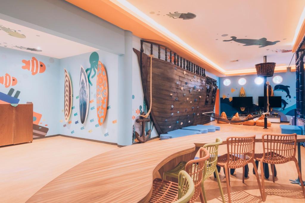 Krāsaina bērnu istaba ar jūras tematiku, dekorēta ar zīmējumiem par zivīm, surfinga dēļiem un jūras dzīvniekiem, kas rada priecīgu un jautru atmosfēru. Iekārtojums ietver koka kuģa imitāciju un ērtas sēdvietas.