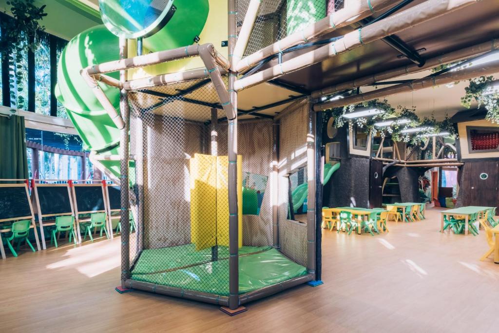 Bērnu rotaļu telpa ar zaļām slidkalniņām, drošības žogu un dzeltenām sienām, kur bērni var spēlēties un mācīties. Apkārt ir zaļi galdi un krēsli.