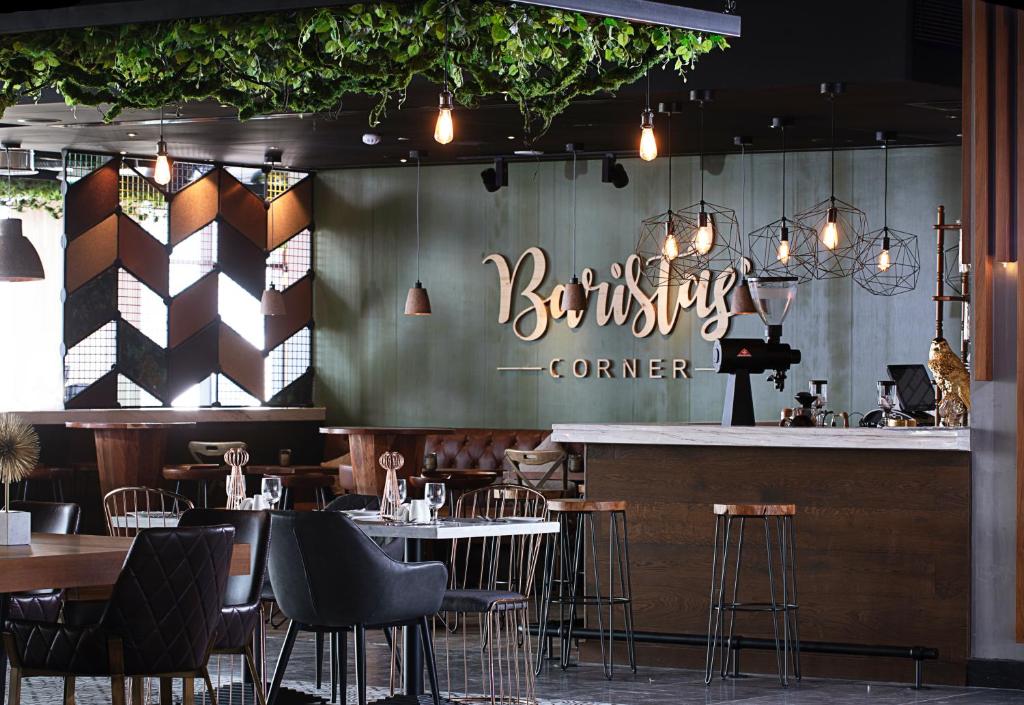 Mūsdienīgs kafejnīcas interjers ar elegantu dizainu, kas ietver zaļus augus, stilīgas gaismas un koka elementus. Uz sienas redzams nosaukums "Baristas Corner".