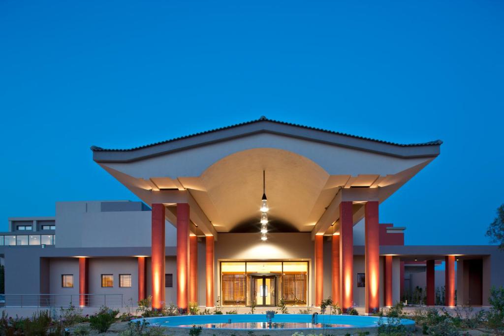 Mūsdienīga arhitektūra ar izteiksmīgu jumtu un sarkaniem stabiem, kas izgaismoti vakarā. Apkārtne ir apzaļumota un mierīga, radot viesmīlīgu atmosfēru.