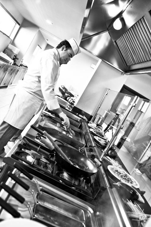 Šefpavārs strādā modernā virtuvē, gatavojot ēdienus uz plīts, kamēr apkārt ir dažādi virtuves piederumi un sastāvdaļas.