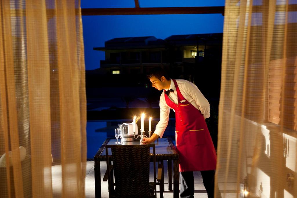 Vakarā apgaismotā terasē, viesmīlis, tērpies rozā priekšautā, kārto galdu ar svečturiem un traukiem. Apgaismojums rada romantisku atmosfēru.